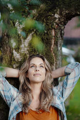 Thoughtful blond woman with hands behind head in front of tree trunk at backyard - JOSEF08209