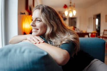 Smiling blond woman leaning on sofa in living room at home - JOSEF08217