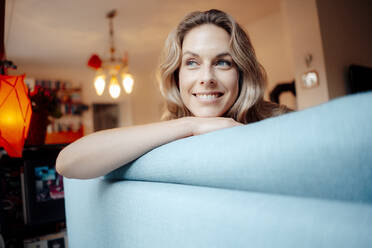Happy blond woman sitting on sofa in living room at home - JOSEF08218