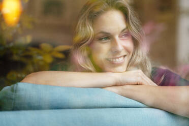 Beautiful smiling woman with blond hair leaning on sofa at home - JOSEF08220