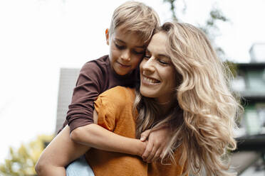 Happy blond woman giving piggyback ride to son at backyard - JOSEF08229