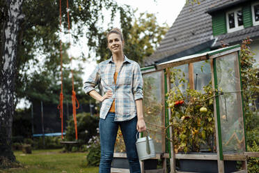 Smiling woman with hand on hip holding watering can in backyard - JOSEF08236