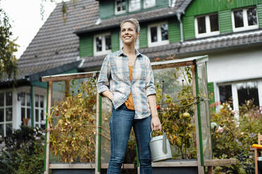 Smiling holding watering can standing with hand in pocket at backyard - JOSEF08237