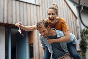 Smiling man giving piggyback ride to cheerful woman holding house key at backyard - JOSEF08249