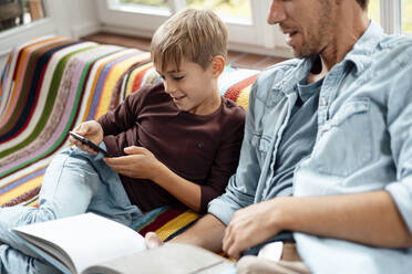 Mann mit Buch, der seinen Sohn bei der Benutzung eines Smartphones beobachtet, sitzt auf dem Sofa zu Hause - JOSEF08253