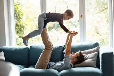 Man holding hands lifting son on legs lying on sofa at home - JOSEF08255