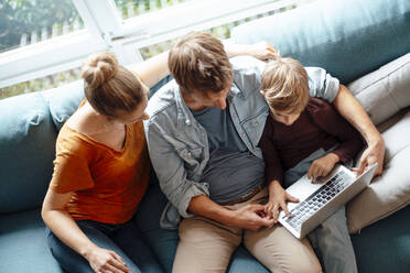 Boy using laptop sitting with parents on sofa in living room - JOSEF08260