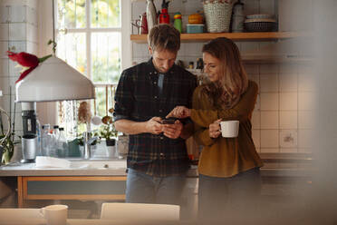 Woman holding coffee cup pointing at smart phone used by boyfriend standing in kitchen - JOSEF08269