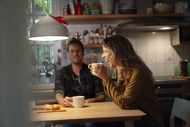 Fröhliche Frau mit Kaffeetasse sitzt mit ihrem Freund am Esstisch in der Küche - JOSEF08270