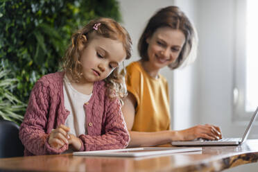 Daughter using tablet PC by smiling mother working at home office - DIGF17753