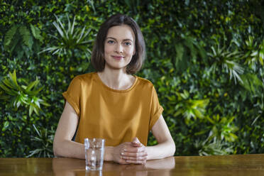 Smiling woman with hands clasped sitting by table at wall with plants - DIGF17765