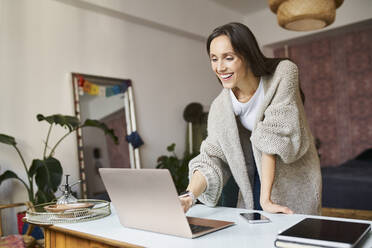 Happy freelancer using laptop leaning on desk at home - BSZF01989