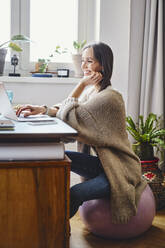 Happy woman sitting on fitness ball using laptop at desk working from home - BSZF01994