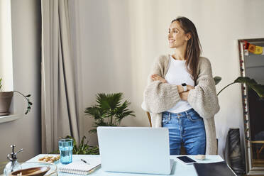 Smiling woman standing with arms crossed by desk at home - BSZF02008