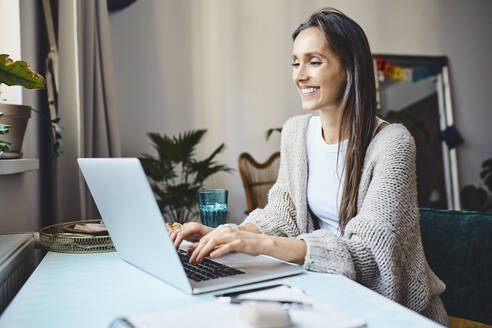 Glückliche Freiberuflerin mit langen Haaren arbeitet zu Hause am Laptop - BSZF02011