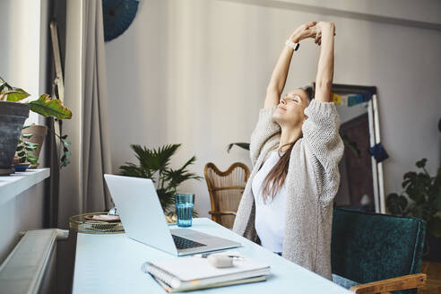 Freelancer with eyes closed stretching arms sitting at desk - BSZF02013