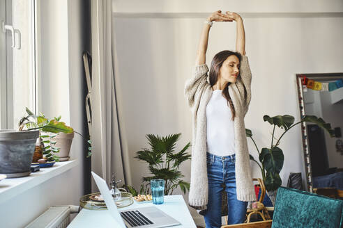 Freelancer stretching arms standing by desk at home office - BSZF02014