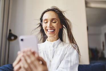Glückliche Frau mit zerzaustem Haar hält Smartphone zu Hause - BSZF02019