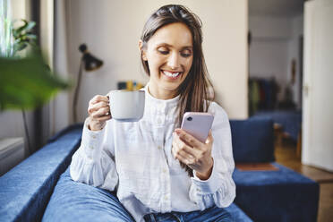 Smiling woman with coffee cup using smart phone sitting on sofa at home - BSZF02020