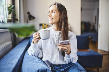 Smiling woman with coffee cup and smart phone sitting on sofa at home - BSZF02021