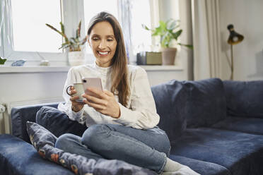 Happy woman surfing net through smart phone sitting on sofa at home - BSZF02022