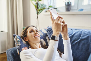 Lächelnde Frau, die ein Smartphone benutzt und zu Hause auf dem Sofa liegt - BSZF02024