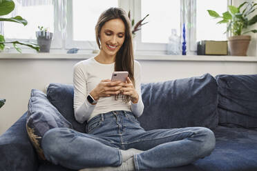 Lächelnde Frau, die ein Mobiltelefon benutzt und im Schneidersitz auf dem Sofa zu Hause sitzt - BSZF02026