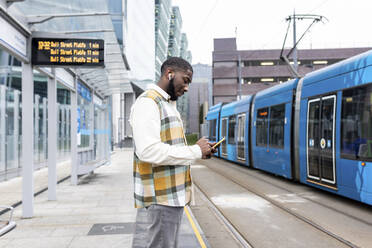 Mann mit drahtlosen In-Ear-Kopfhörern, der an einer Straßenbahnhaltestelle ein Mobiltelefon benutzt - WPEF05863