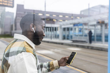 Mann hört Musik über kabellose In-Ear-Kopfhörer und benutzt ein Mobiltelefon an einer Straßenbahnhaltestelle - WPEF05865