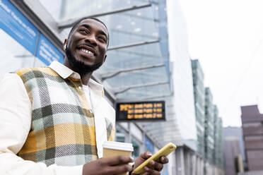 Smiling man holding disposable cup and mobile phone at tram station - WPEF05869