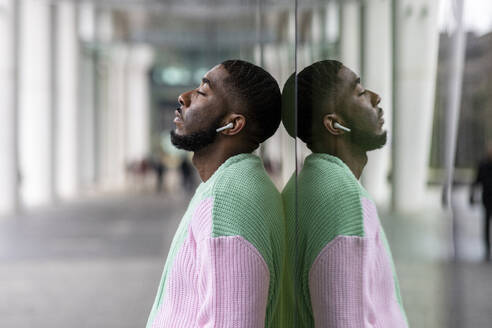 Man with eyes closed listening music through wireless in-ear headphones leaning on glass wall - WPEF05882