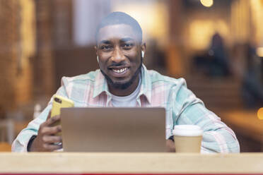 Glücklicher Mann mit Laptop und Smartphone im Café sitzend durch Glas gesehen - WPEF05899