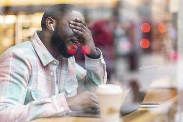 Worried freelancer with laptop sitting in cafe seen through glass - WPEF05903