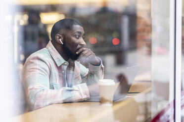 Nachdenklicher Freiberufler mit Laptop, der in einem Café sitzt, gesehen durch Glas - WPEF05904