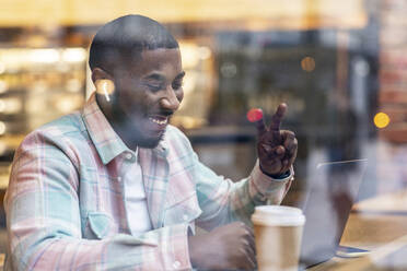 Happy freelancer gesturing peace sign on video call through laptop at cafe seen through glass - WPEF05906