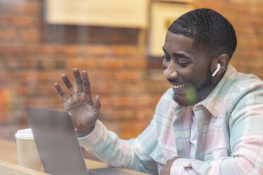 Happy freelancer waving hand on video call through laptop sitting at cafe - WPEF05907