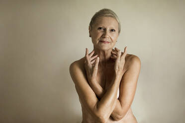 Smiling topless senior woman covering breasts against white background - LLUF00468