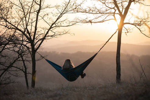 Blonde girl sitting in hammock at sunset - CAVF95920