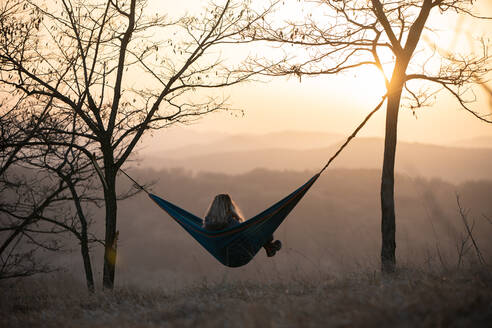 Blondes Mädchen sitzt in der Hängematte bei Sonnenuntergang - CAVF95920