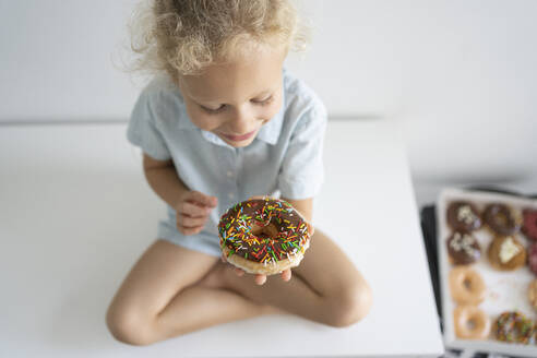 Smiling girl with doughnut sitting cross-legged on table - SVKF00045