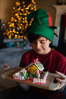 Fröhlicher Junge mit Elfenmütze betrachtet ein Lebkuchenhaus zur Weihnachtszeit. - CAVF95951