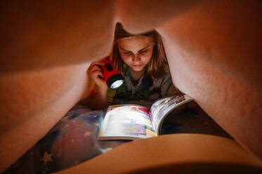 Junges Mädchen liest Buch unter rosa Decke mit Taschenlampe - CAVF95953