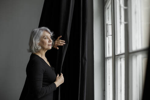 Ältere Frau steht vor einem Vorhang und schaut durch ein Fenster zu Hause - LLUF00473
