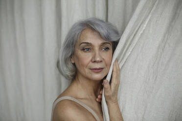 Nachdenkliche ältere Frau mit grauem Haar, die den Vorhang zu Hause berührt - LLUF00484