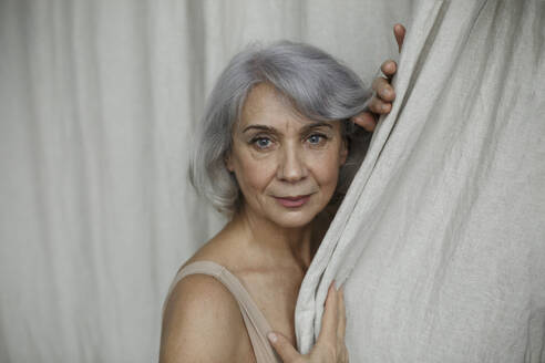 Ältere Frau mit grauen Haaren vor einem Vorhang zu Hause - LLUF00485