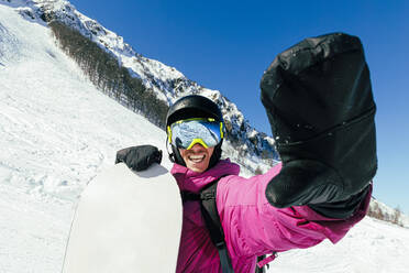 Happy man holding snowboard standing at snowcapped mountain - OMIF00755
