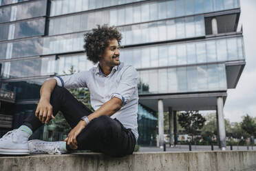 Smiling young man sitting on ledge in front of modern building - MFF08926