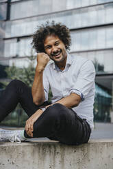 Happy young man with clenched fist sitting on ledge in front of building - MFF08928