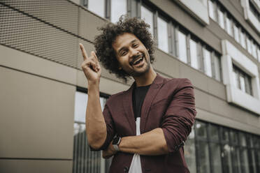 Happy young man with curly hair pointing in front of building - MFF08936