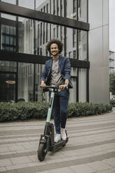 Glücklicher junger Mann fährt mit einem elektrischen Roller auf dem Fußweg vor einem modernen Gebäude - MFF08953